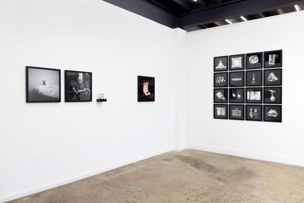 Installation view of “Evelyn Sosa: No Place is Far Away” at Mahara+Co., Miami. From left, portrait of Armando, a still life of a photo of his mother, and the original photo of his mother placed on a black shelf, a color image of a red jewelry box with two dried umbilical cord ends, and an arrangement of black and white images of small mementos brought by Cuban immigrants to America. (Photo courtesy of Mahara+Co.).